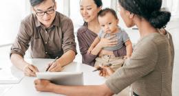 A woman is presenting a document to a man who is signing it, while another woman holds a baby.
